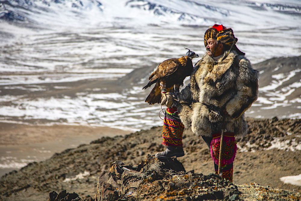 A proud eagle hunter holding his golden eagle while standing on a rocky cliffside overlooking a snowy landscape