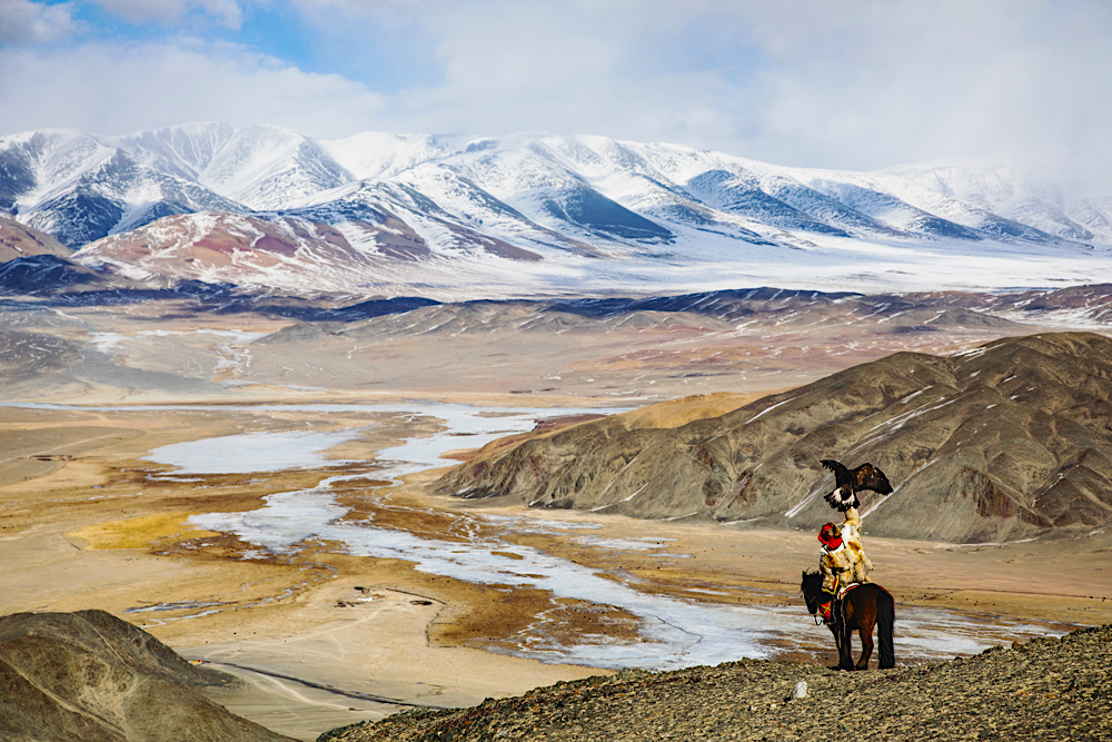 An eagle hunter hunting with his golden eagle on horseback overlooking a cliff trying to find prey